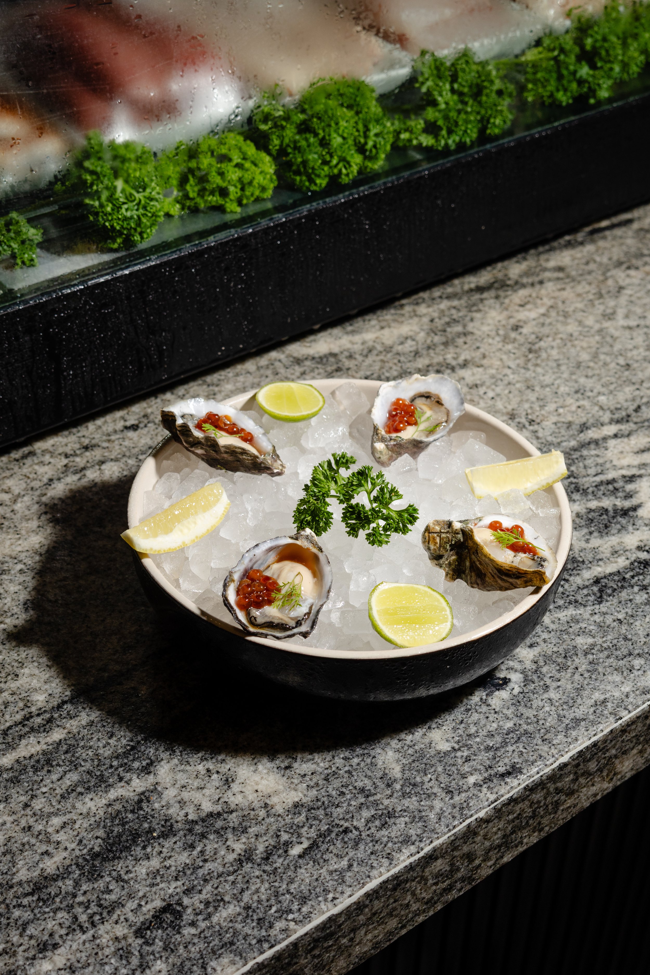 Oysters on a bed of ice with lime wedges and parsley in a white bowl, set on a granite countertop, with a wall-mounted fish display case with greens and fish in the background.