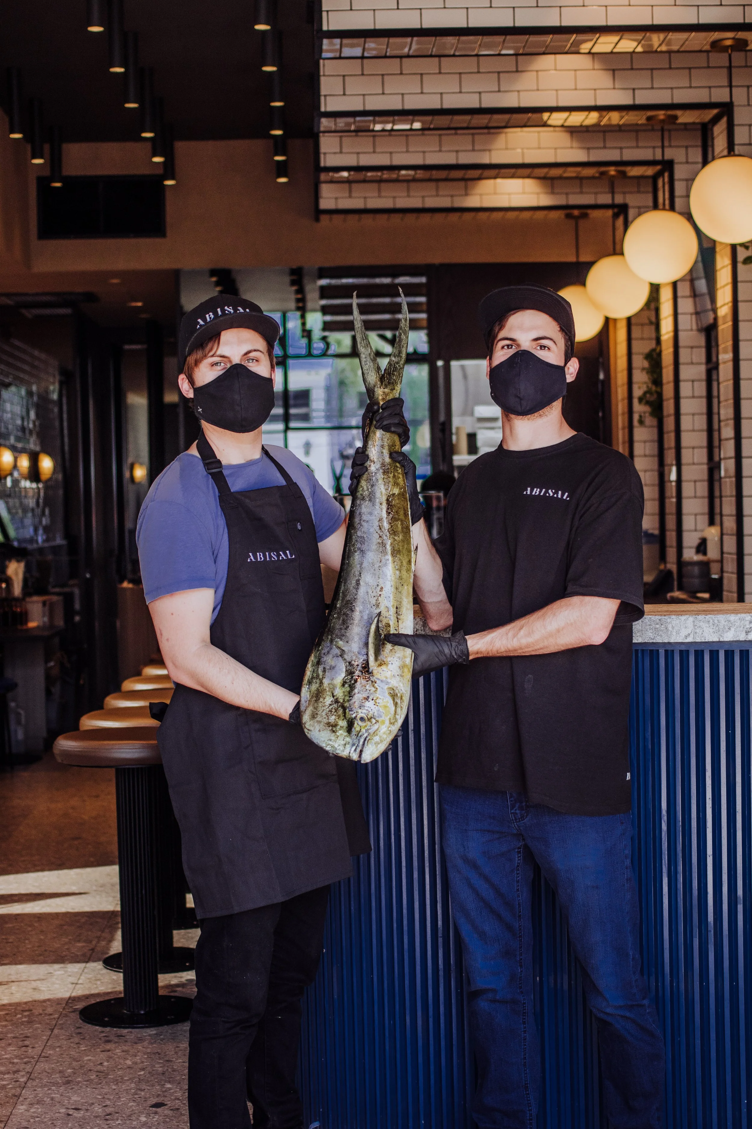 Two young men wearing black masks, black aprons, and black caps, holding a large fish between them inside a restaurant. The man on the left is wearing a blue t-shirt, and the man on the right is wearing a black t-shirt. They are standing behind a blue counter with a modern interior decor featuring hanging lights and brick walls.