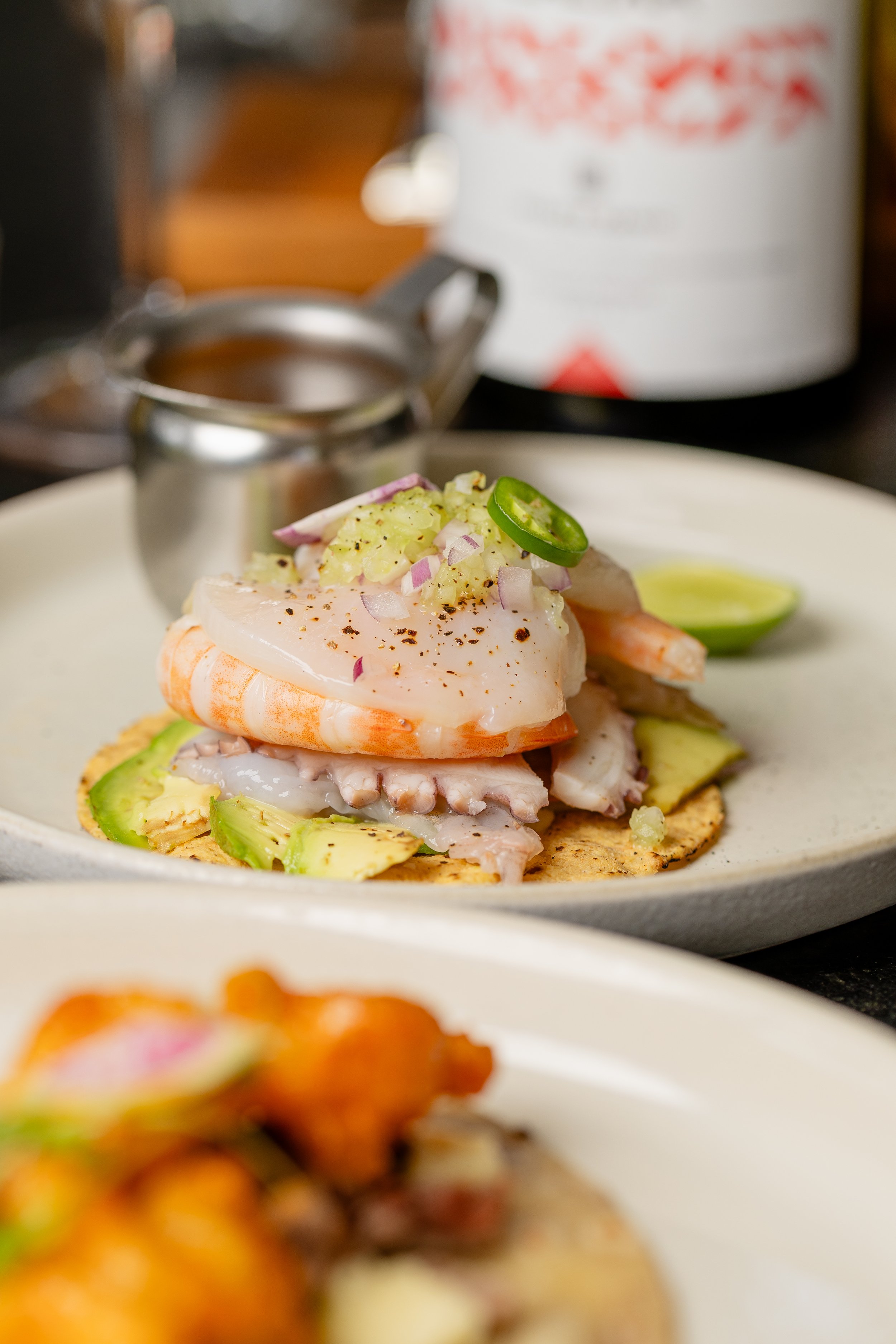 Close-up of a shrimp tostada topped with diced onions, jalapeño slices, and chopped vegetables, served on a white plate with a lime wedge, in a restaurant setting.