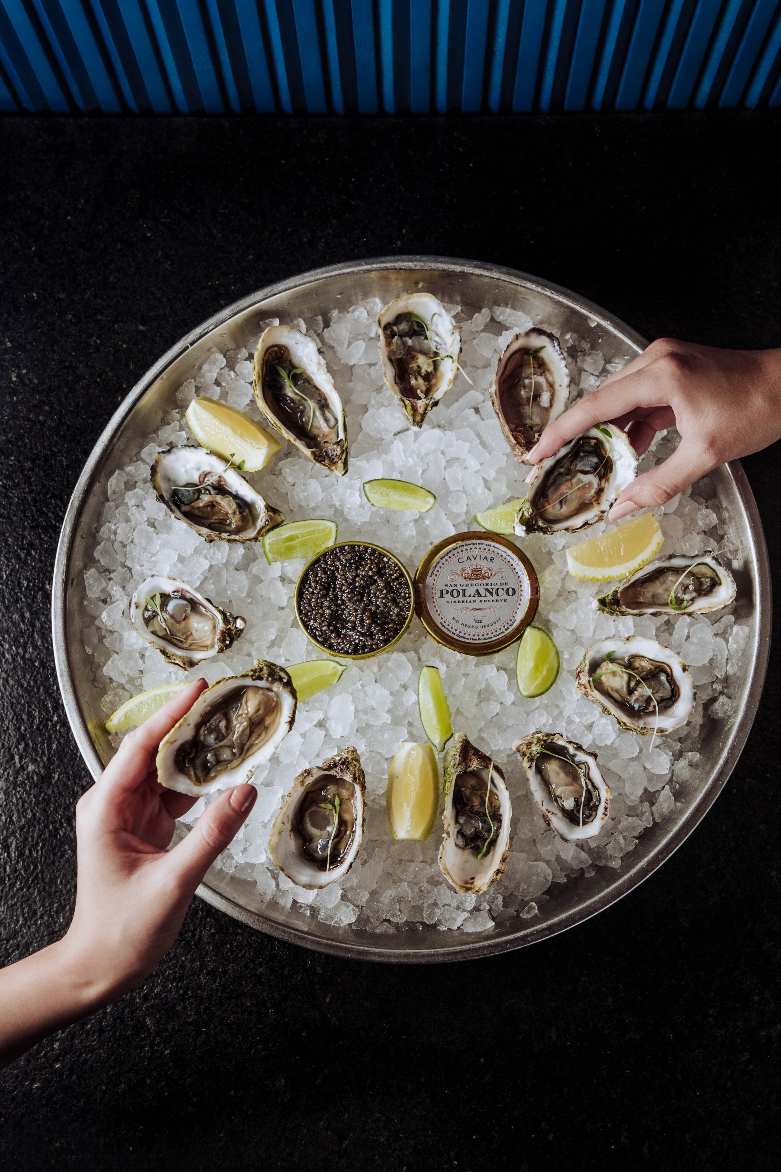 A round metal tray filled with ice and dozens of oysters on the half shell, garnished with small lime wedges and containers of black caviar, on a black countertop.