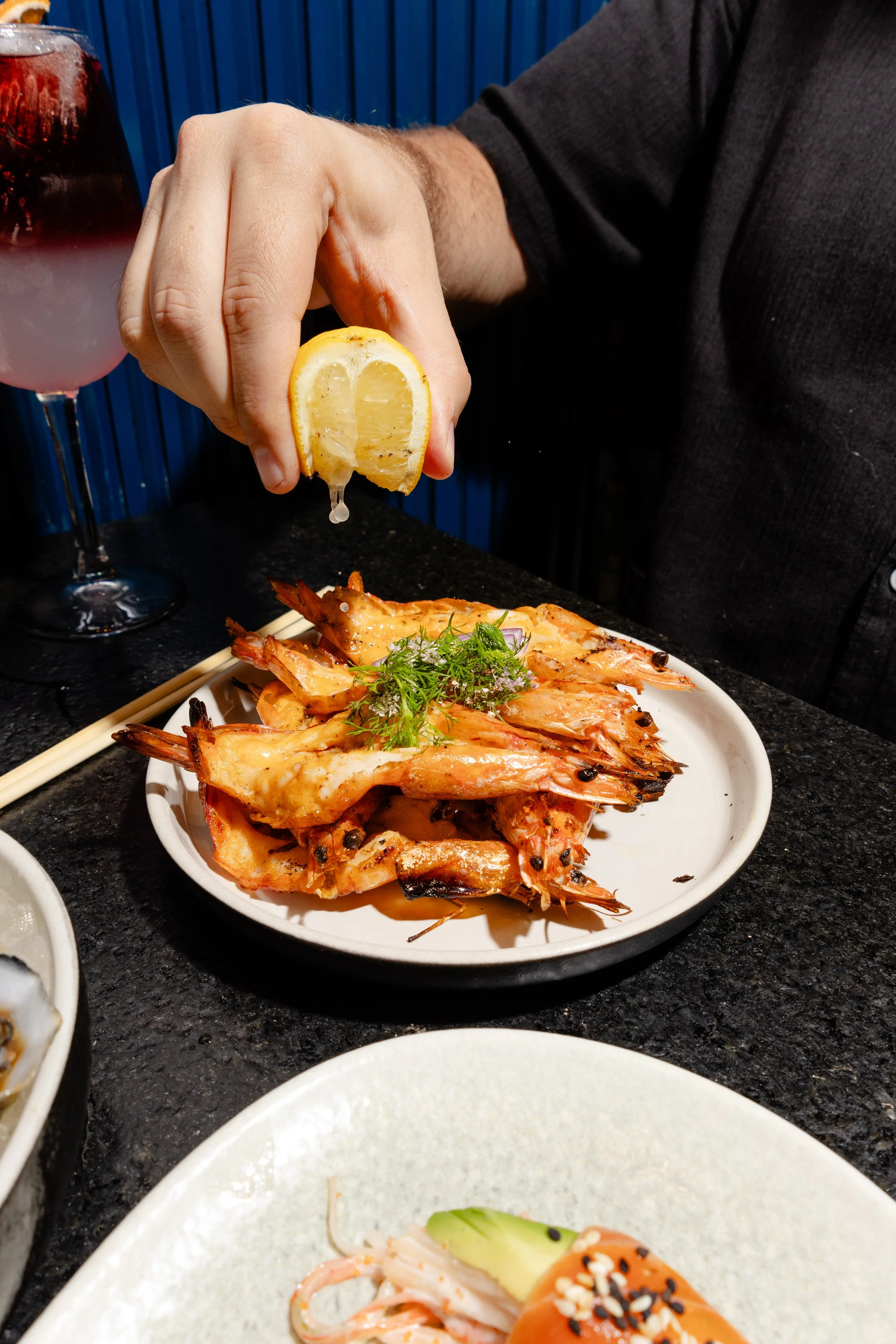 A person squeezing a lemon over a plate of grilled prawns garnished with herbs, with a glass of dark beverage in the background.