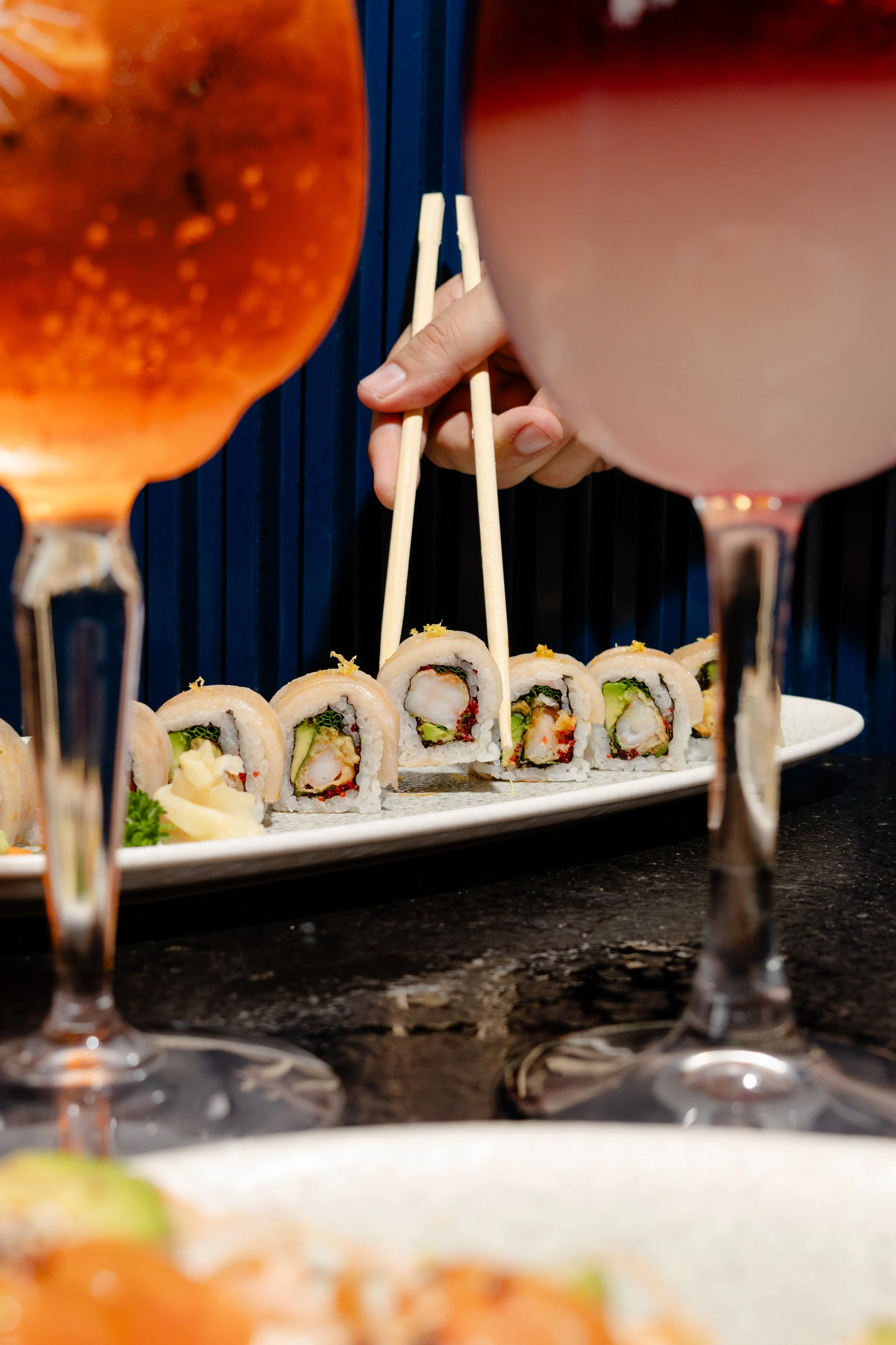 Close-up view of a platter of assorted sushi rolls with various ingredients, garnished with wasabi and pickled ginger, on a dark granite table, with two wine glasses partially visible in the foreground.