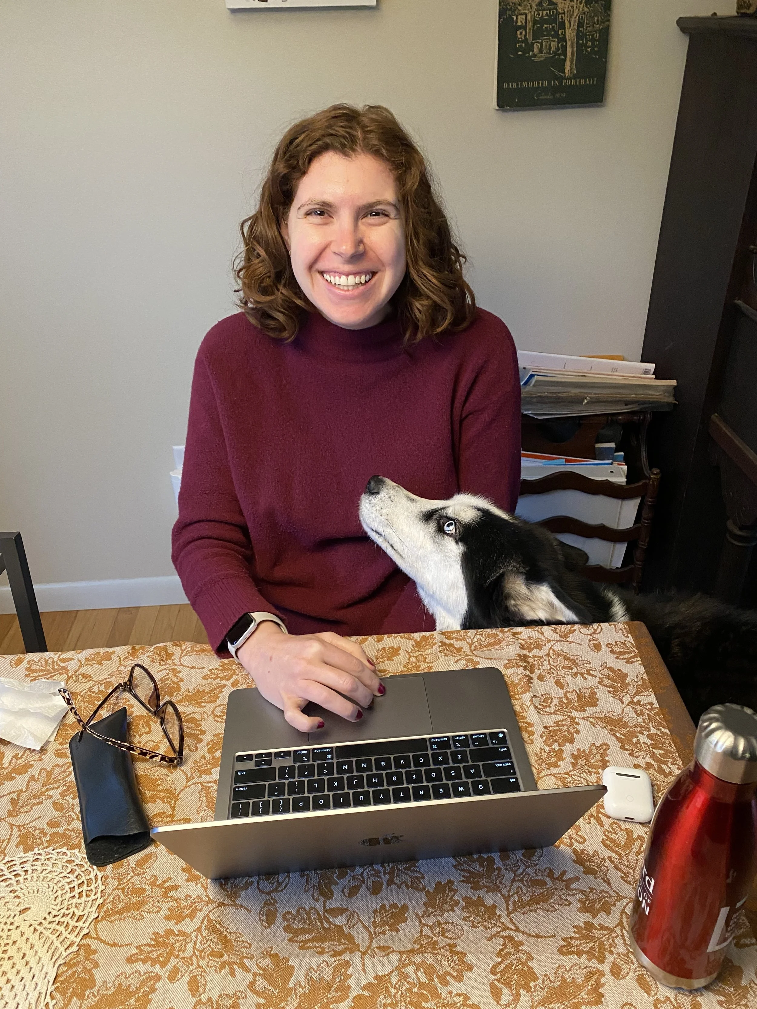 Smiling woman in a burgundy sweater working on a laptop at a dining table with a black-and-white dog beside her.