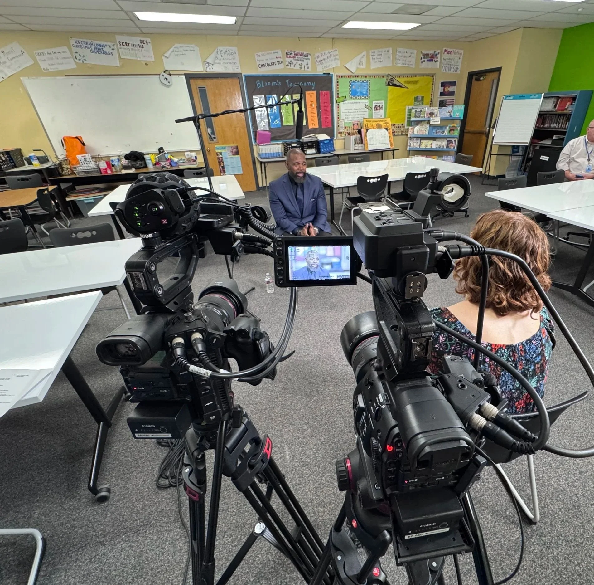 Two professional video cameras film a man seated in a classroom surrounded by educational posters and tables.