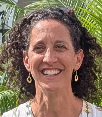 A smiling woman with dark curly hair, wearing earrings, outdoors with green tropical plants in the background.