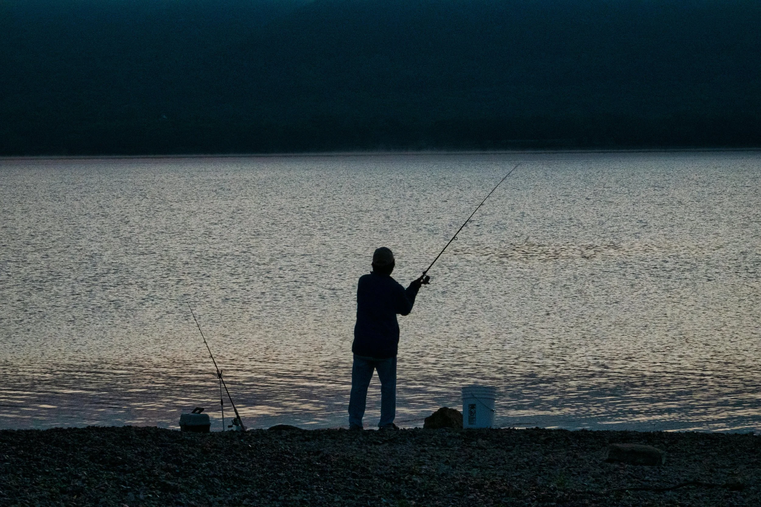 A person fishing at sunset on a calm body of water, with two fishing rods, buckets, and a rocky shoreline.