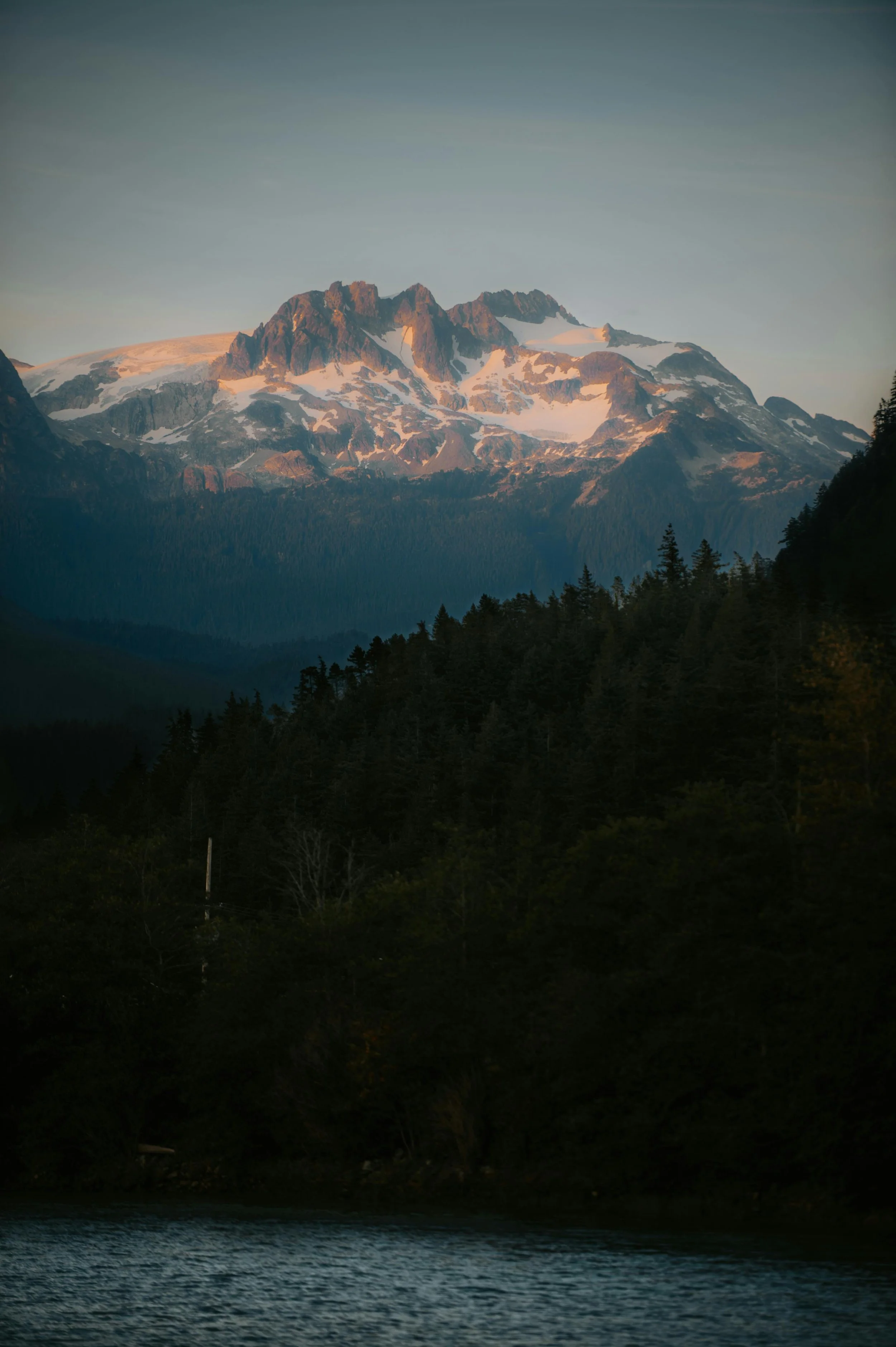 A mountain with snow patches and a forested base, reflected in a body of water in the foreground, under a partially cloudy sky.
