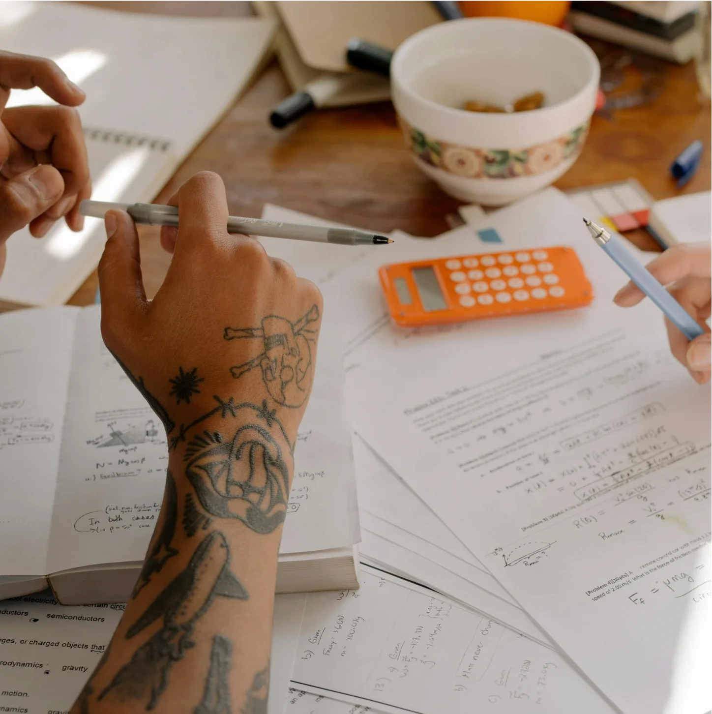 Hands with tattoos working on physics or math homework, surrounded by papers, a calculator, and writing utensils on a cluttered desk.