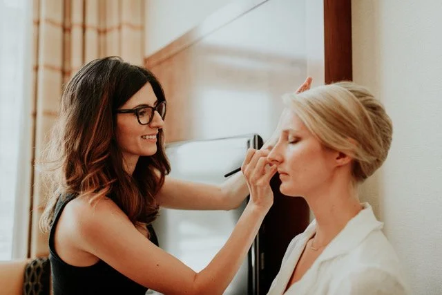 A makeup artist applying makeup to a woman seated against a wall in a room.
