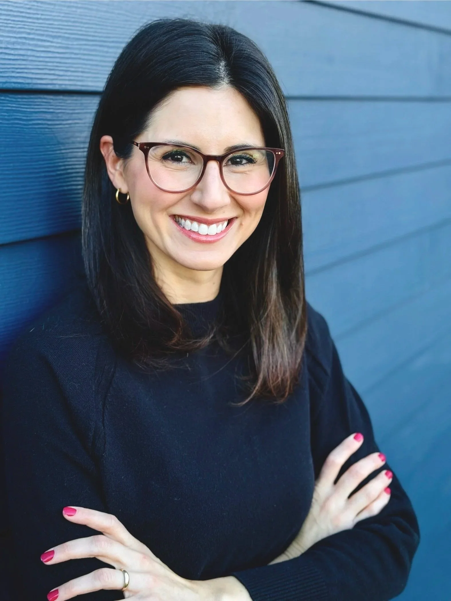 A woman with long dark hair, wearing glasses, smiling, and crossing her arms with nails painted pink, standing against a blue wall.