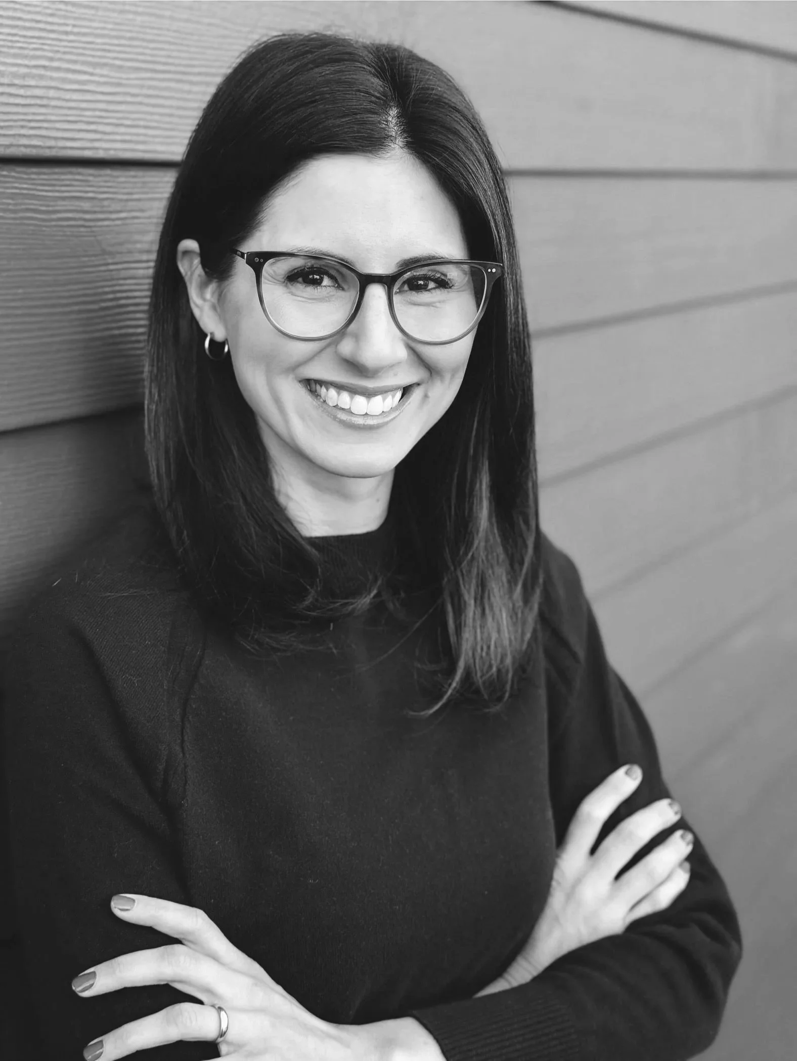 Black and white photo of a woman with glasses, smiling, wearing a dark sweater, with her hands crossed on her chest, standing in front of wooden wall panels.
