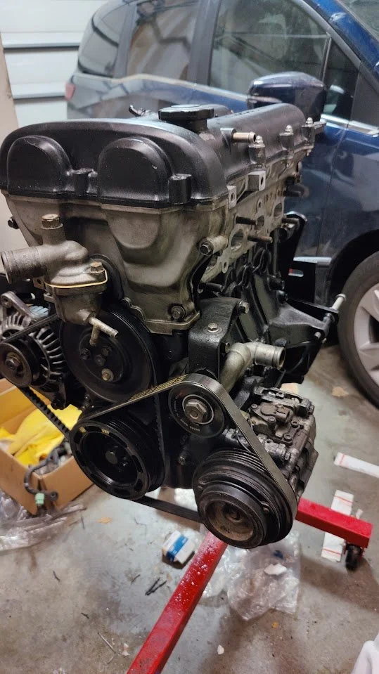 A car engine mounted on a red engine stand in a garage, with other vehicles and tools visible in the background.