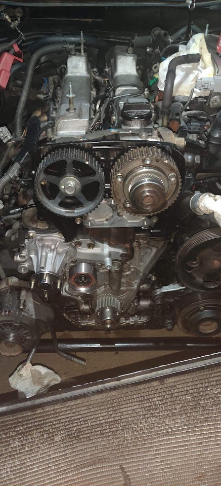 Inside a car engine bay, showing the timing belt, camshaft gears, and engine components with some parts disconnected.