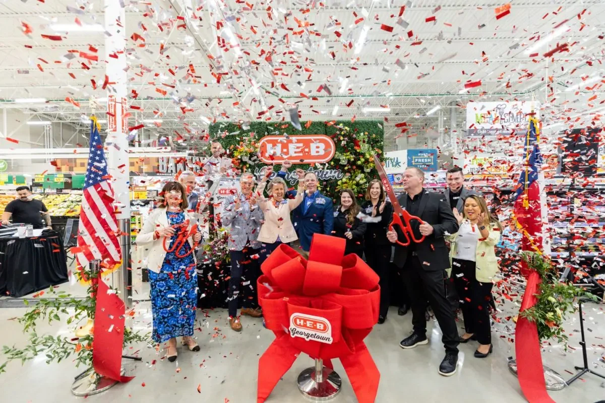 Group of people celebrating grand opening of a H-E-B store with confetti, flag, big scissors, and decorative ribbon, in a grocery store