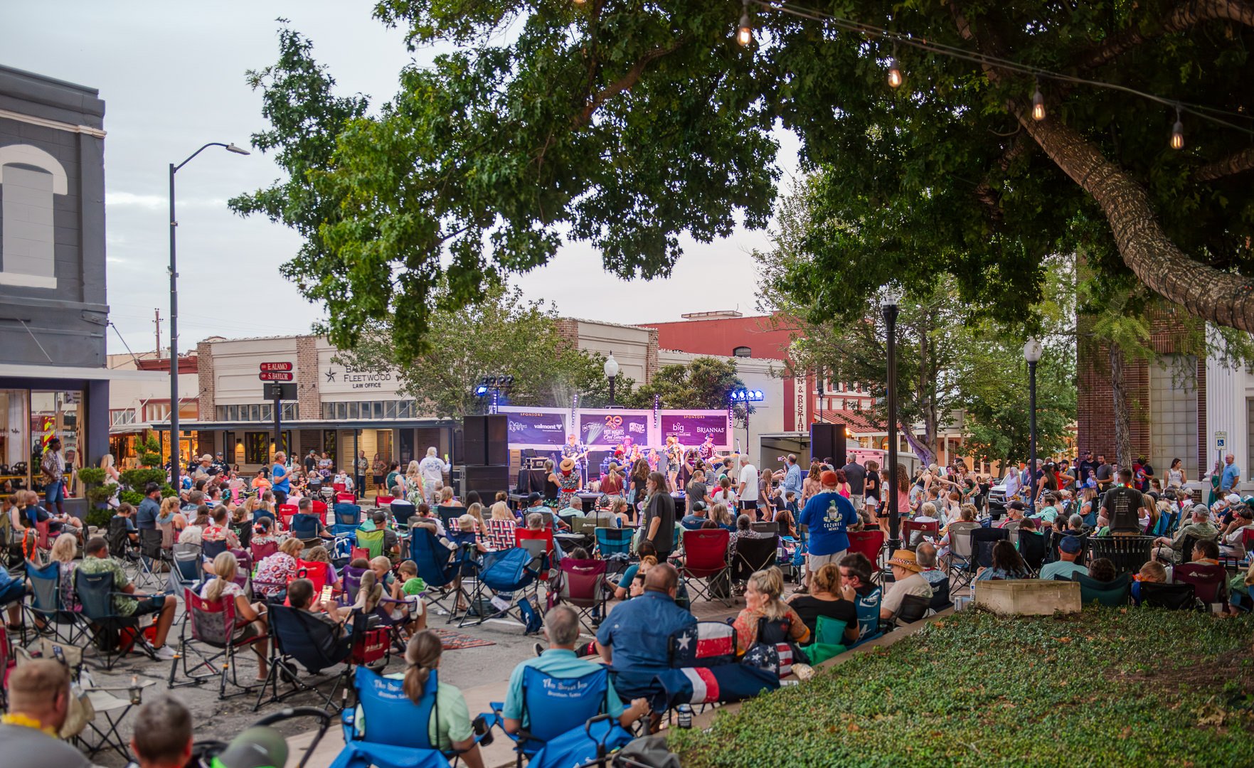 Outdoor concert in a town square with a band performing on stage, surrounded by a large crowd sitting on chairs and standing, some people wearing cowboy hats, trees, string lights, and city buildings in the background.