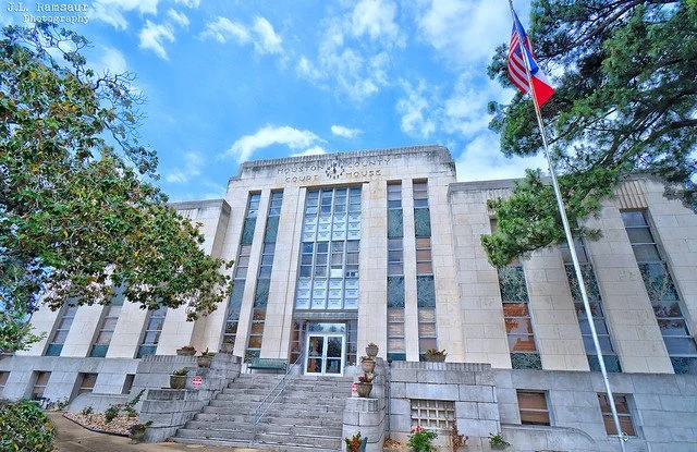View of a large courthouse building with steps leading up to the entrance, surrounded by trees, with an American flag on a flagpole in front, and a blue sky with some clouds overhead.