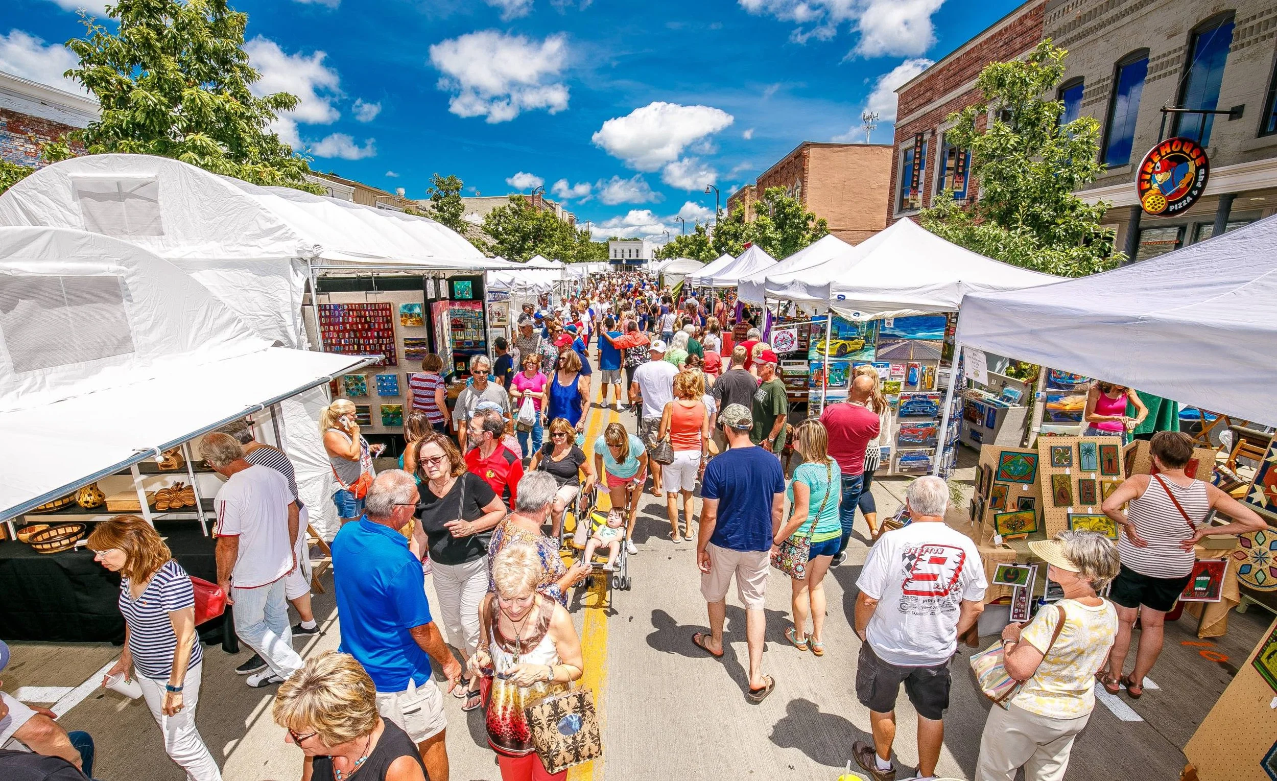 A crowded outdoor street fair with numerous white tents and art and craft booths. People of various ages are walking and shopping on a sunny day with a partly cloudy sky. Some trees and brick buildings are visible in the background.