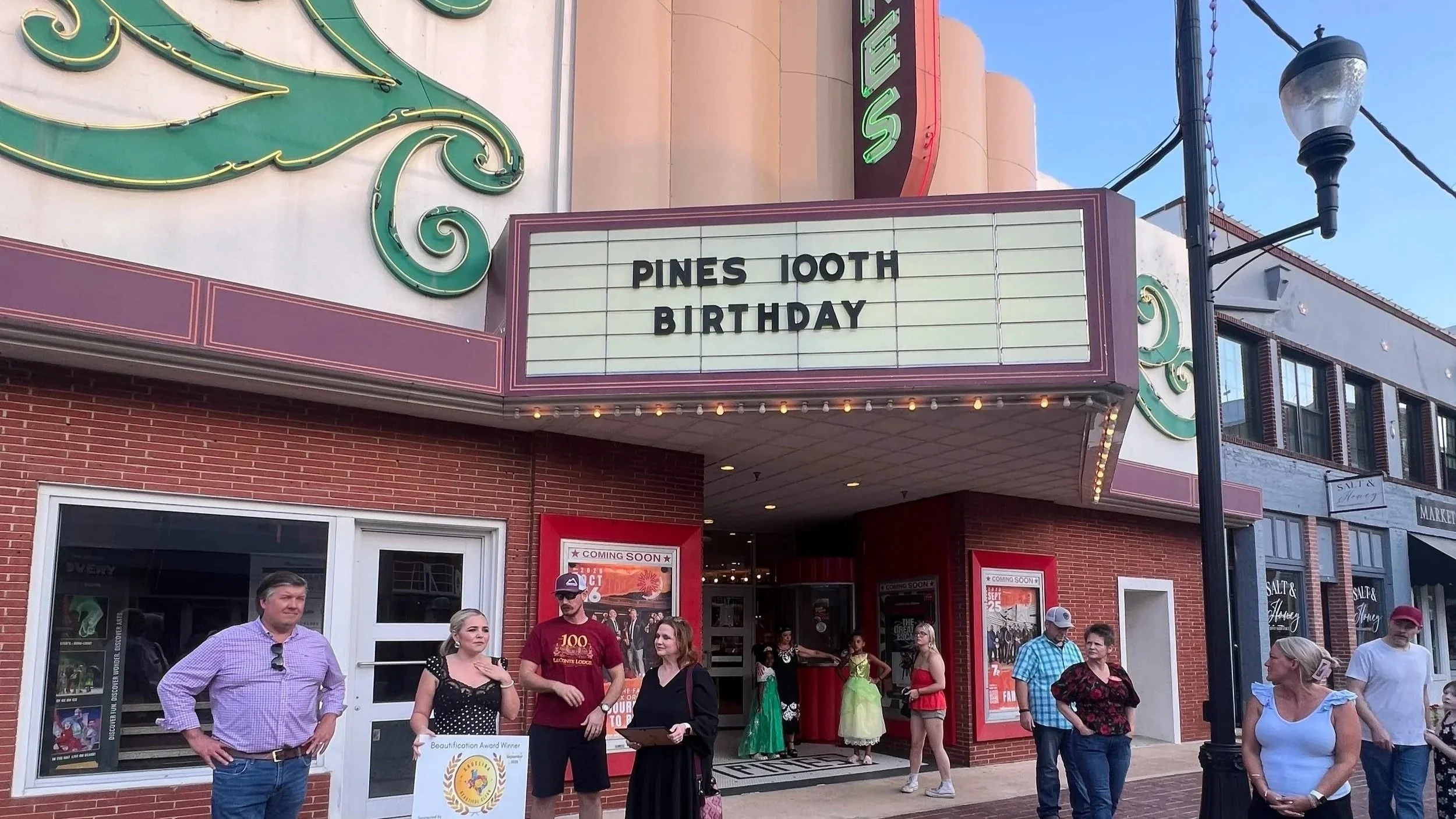People standing in front of a theater marquee that reads 'PINES 100TH BIRTHDAY' with some theater posters and actors in costume visible inside.