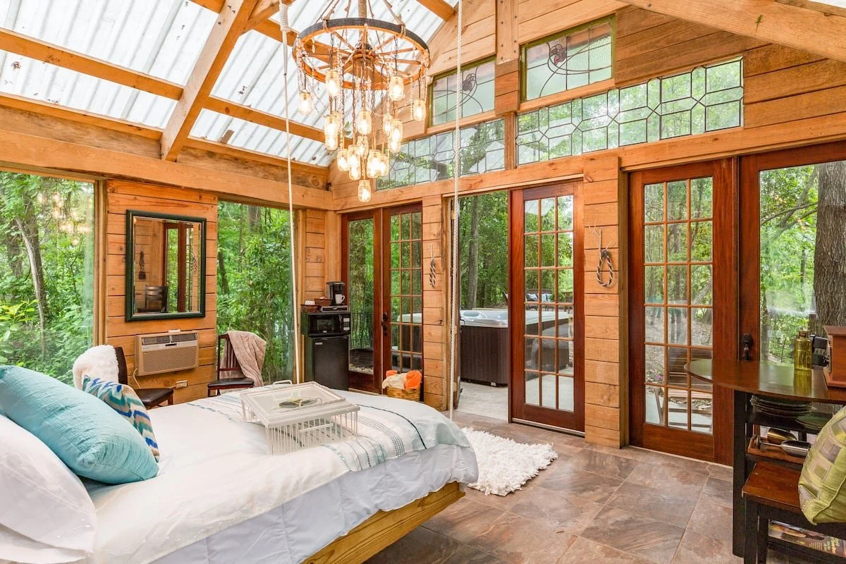 A cozy bedroom with wooden walls and large glass doors and windows showing green trees outside. There is a bed with pillows and a tray, a chandelier hanging from the ceiling, and a small outdoor patio visible through the sliding doors.