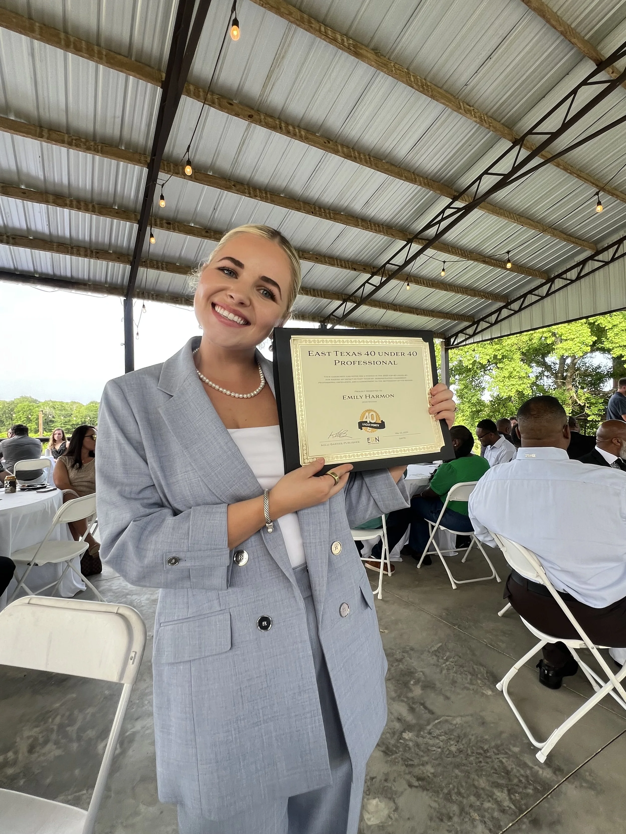 A young woman smiling, wearing a gray blazer and pearl necklace, holding a framed certificate at an outdoor event under a large metal pavilion. The certificate has the title 'East Texas 40 Under 40 Professional' and the woman's name 'Emily Harmon'.