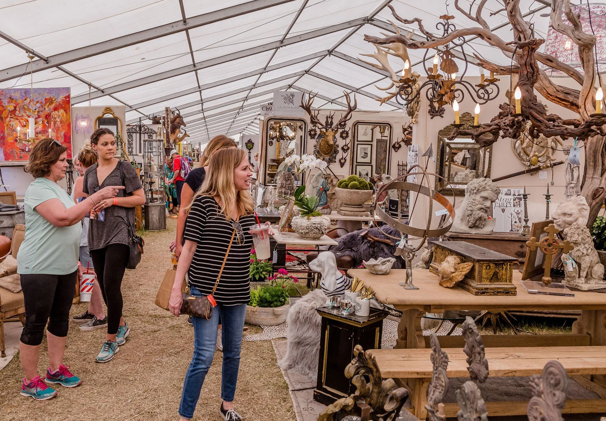 People browsing antique and vintage items at an outdoor market, with displays of sculptures, mirrors, and decorative art pieces inside a tent.