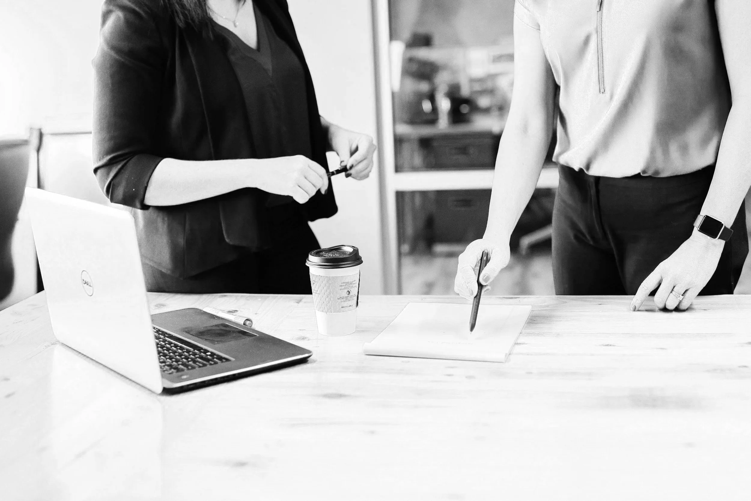 Two women standing at a table, engaged in a discussion or meeting. One woman is holding a pen and pointing to a paper, while the other has her hand on a paper. On the table, there is a laptop, a disposable coffee cup, and some papers.