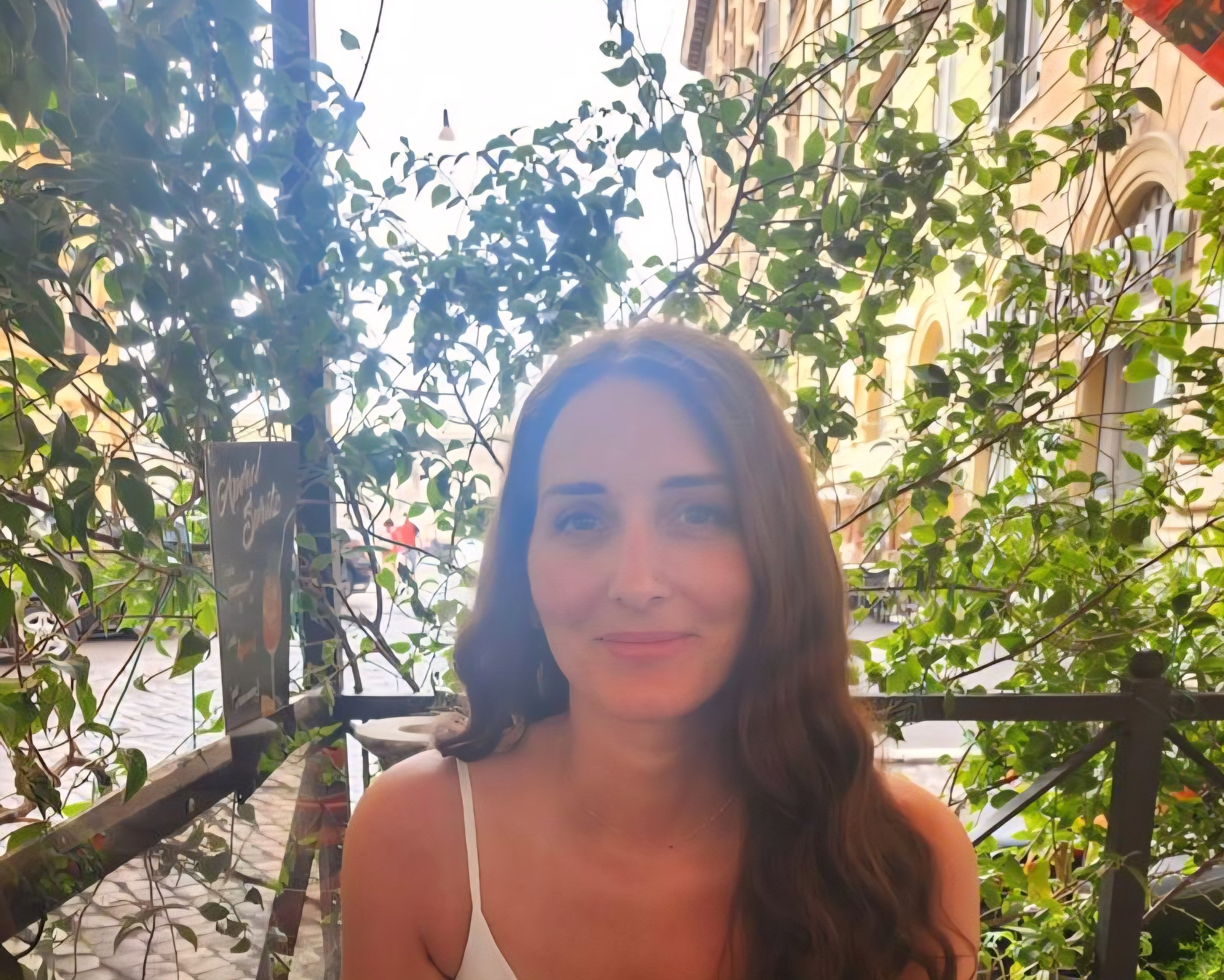Young woman with long brown hair and a white tank top, smiling outdoors behind green leafy plants with a building in the background.