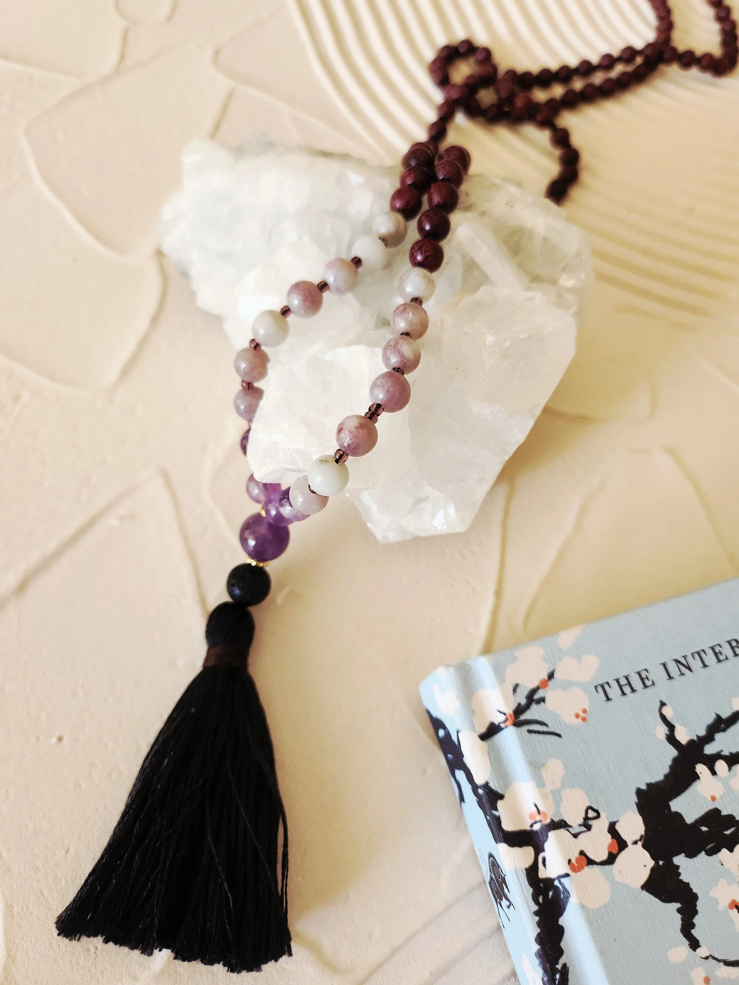A beaded necklace with alternating white and pink beads, a black tassel at the end, and a block of white rough crystal on a light-colored surface with a textured pattern, partially overlapping a book with a black and white tree illustration.