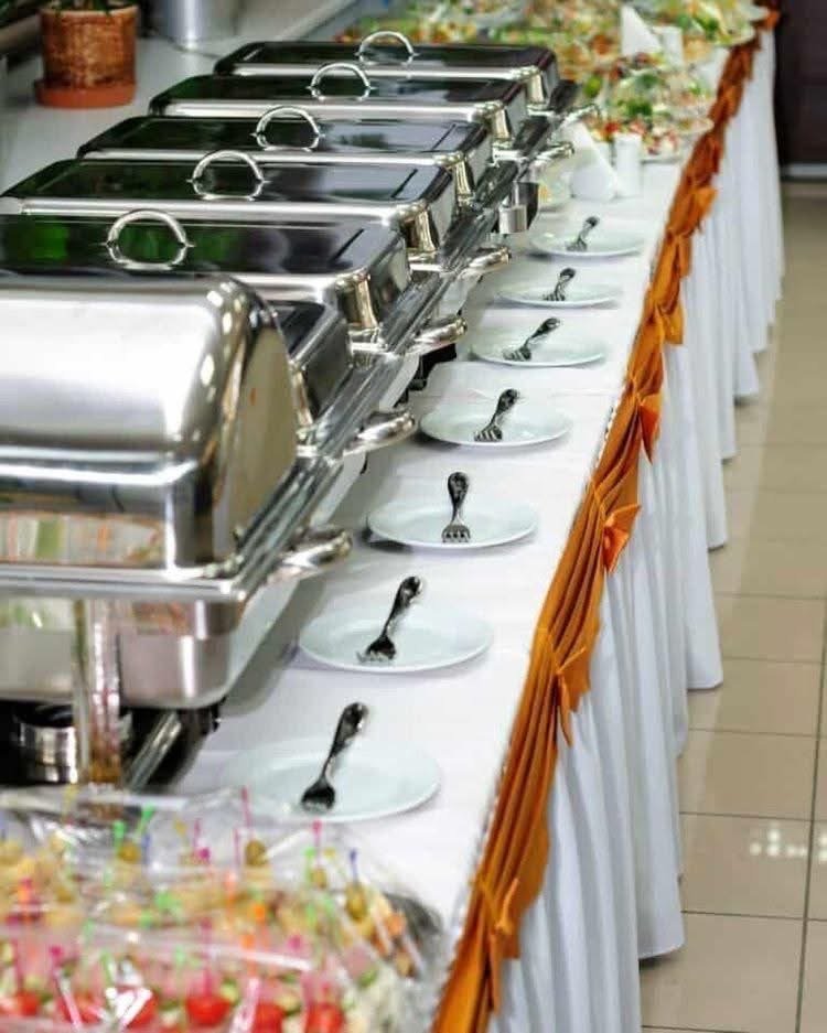 Buffet table with chafing dishes, empty plates with forks arranged in a line, and a nearby section of colorful candies, all decorated with orange ribbons.