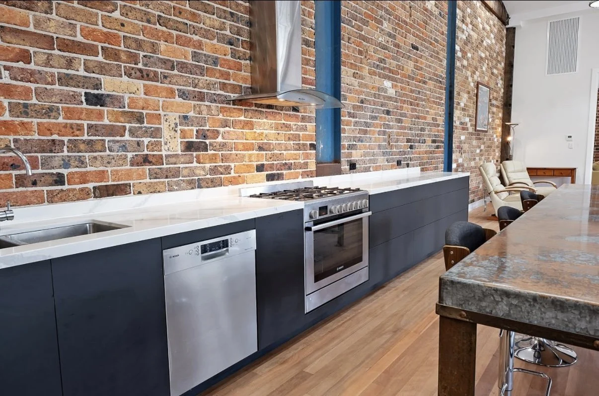Modern kitchen with black cabinets, stainless steel oven, dishwasher, and range hood, exposed brick wall, wooden floor, and a marble-top dining table with chairs.
