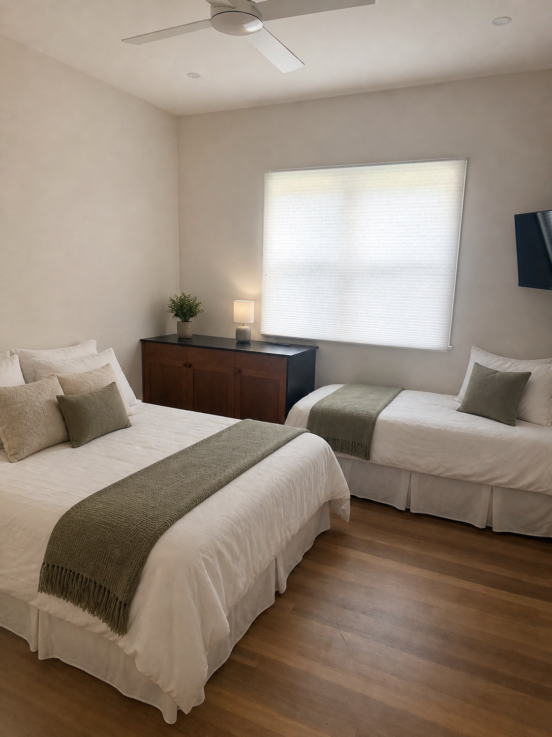 Bedroom with two beds, white bedding, green throws, pillows, wooden floor, window with closed white blinds, side table with lamp and plant, ceiling fan, and wall-mounted TV.