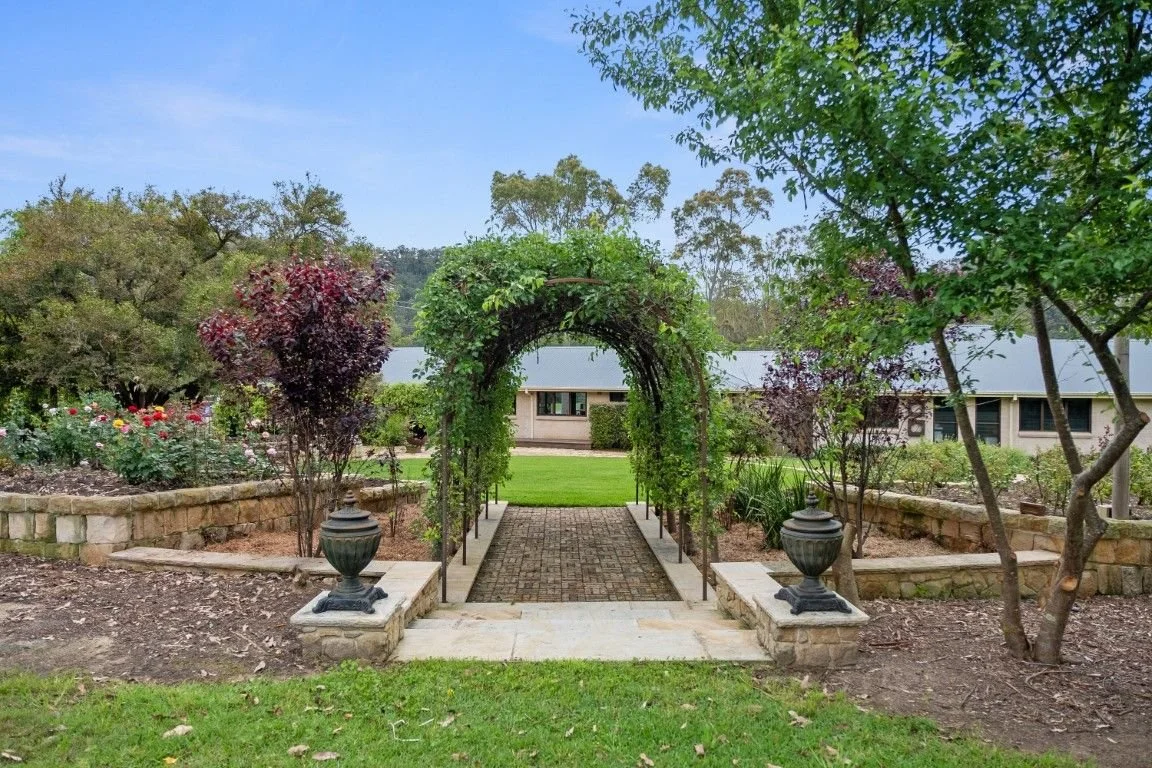 A garden pathway with an archway covered in green vines, flanked by flowerbeds with roses and shrubs, leading toward a building in the background under a blue sky.