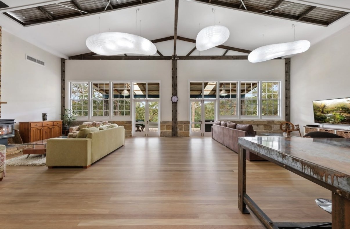 Spacious living room with large windows, modern white ceiling lights, wooden beams, and hardwood floors, furnished with sofas and a television.