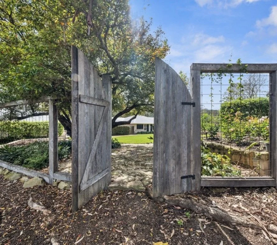 Open wooden gate leading to a backyard garden with a tree and a house in the background under a partly cloudy sky.