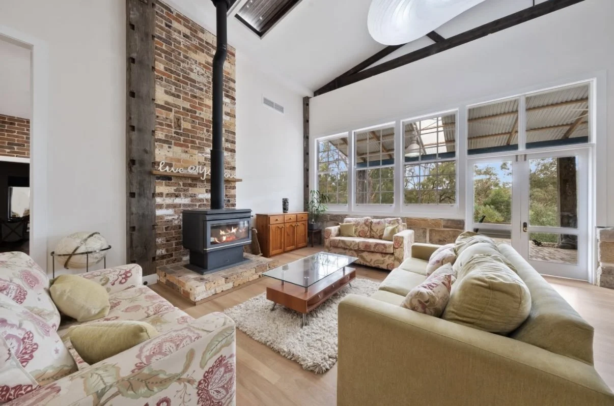 Living room with large windows, yellow and floral patterned sofas, a brick fireplace with a stove, a wooden cabinet, and a glass coffee table.