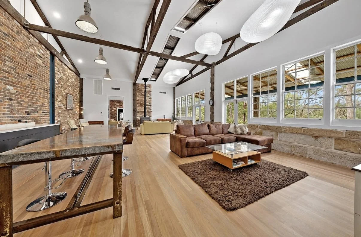 Spacious loft-style living room with exposed brick walls, large windows, wooden beams, and modern furniture including a brown sectional sofa, a glass coffee table, and bar stools at a high countertop.