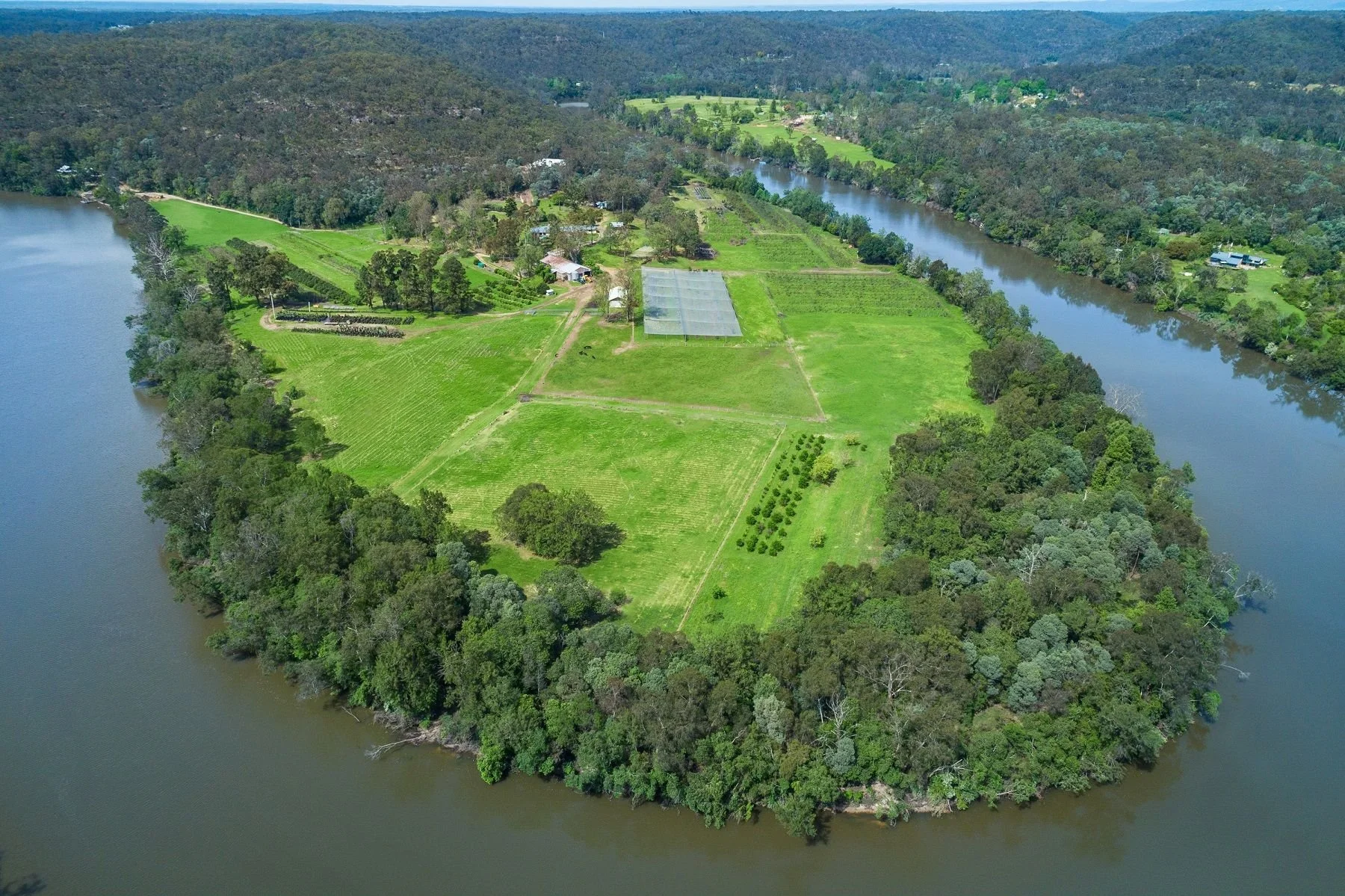 Aerial view of a large island with green fields, trees, a few buildings, and a greenhouse, surrounded by a river in a lush landscape.