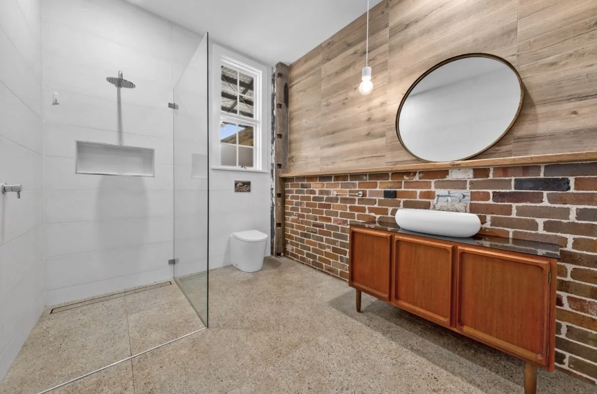 Modern bathroom with white tiled shower area, glass door, white toilet, a small window, brick wall, wooden wall panel, oval mirror, vessel sink on wooden vanity, exposed brick and wood accents, and a pendant light.