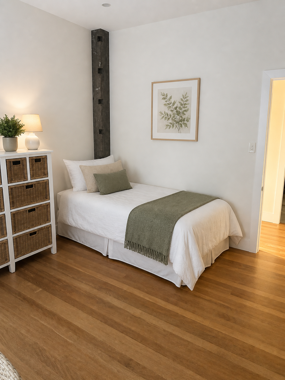 A cozy bedroom with a white bed, green throw blanket and pillows, a white dresser with wicker baskets, a lamp, a potted plant, and a framed botanical print on the wall.