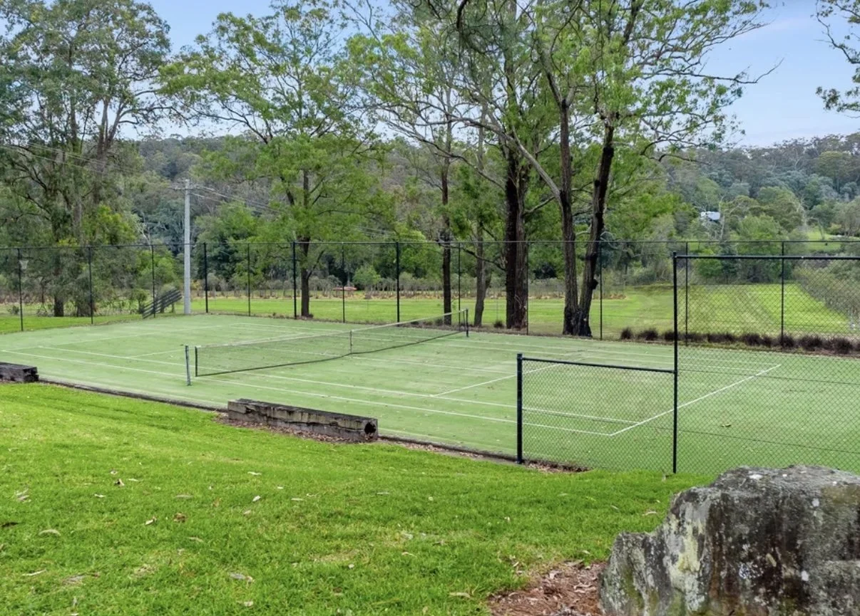 Empty outdoor tennis court with green surface and chain-link fencing, surrounded by trees and grassy area.