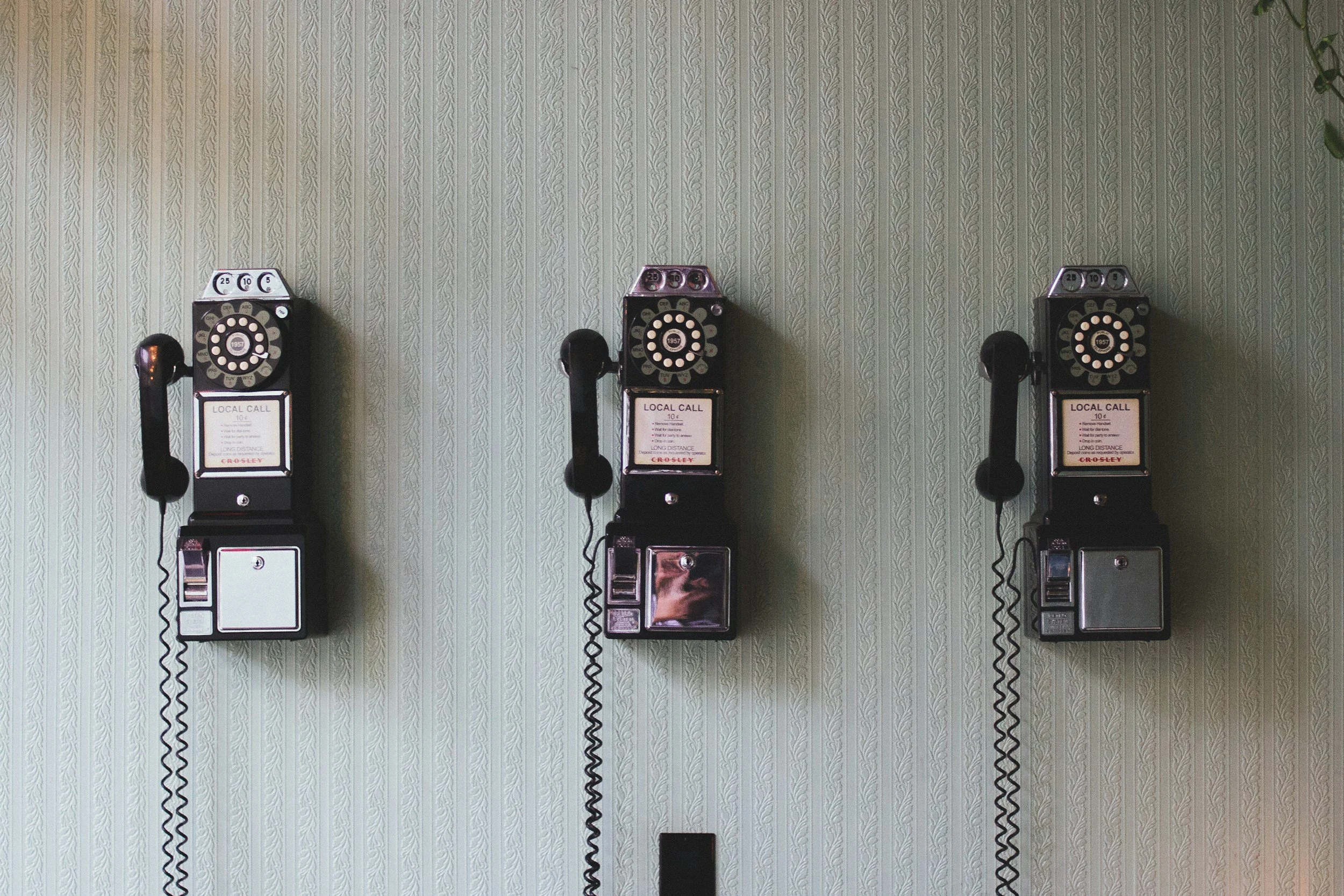 Three vintage black rotary dial payphones mounted on a wall with green patterned wallpaper.
