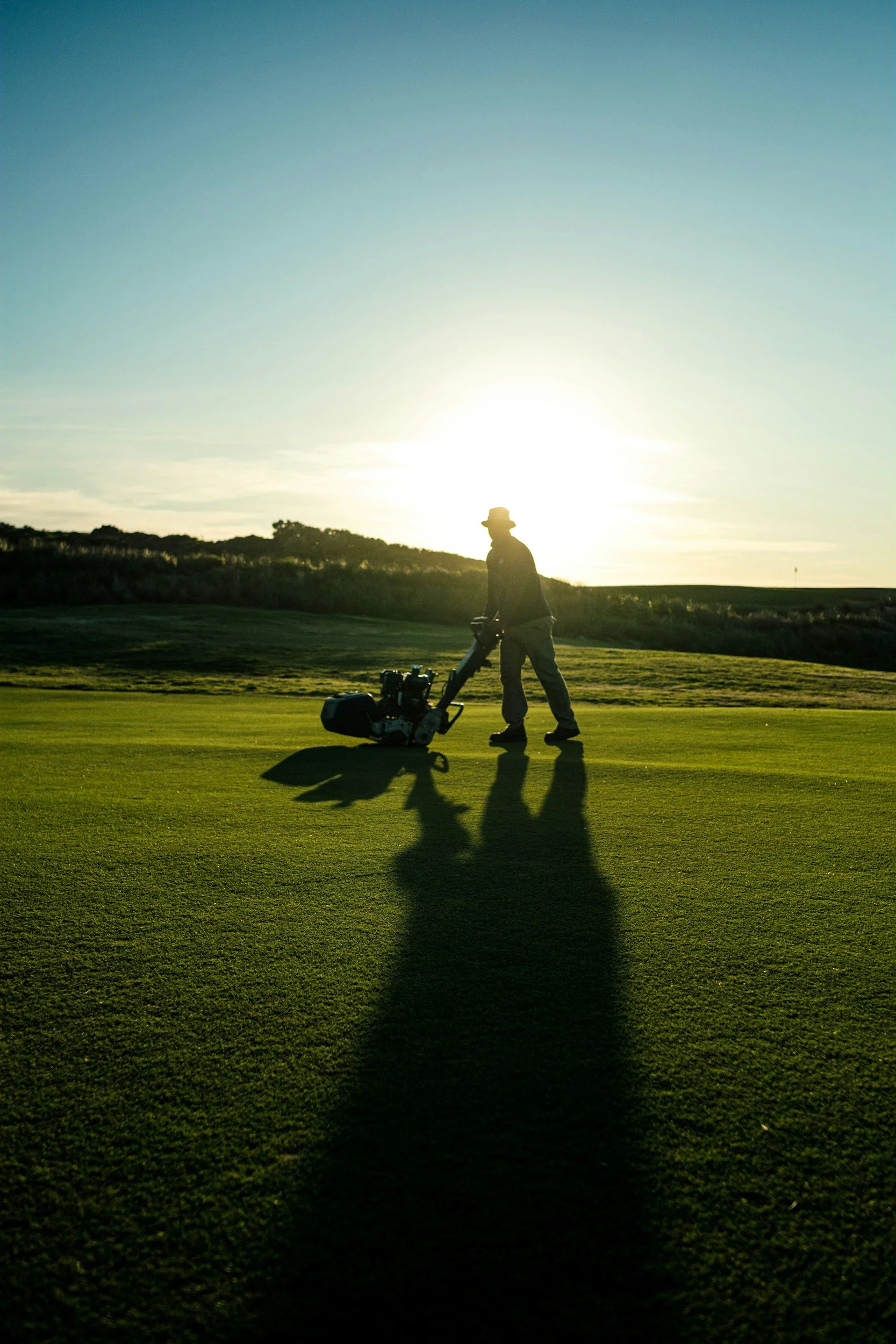 A person operating a lawn mower on a golf course at sunset, with long shadows cast on the grass.