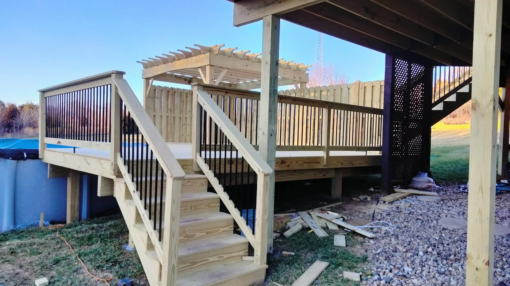 Wooden deck under construction, with stairs and black metal railings, attached to a house, with gravel and construction debris below.