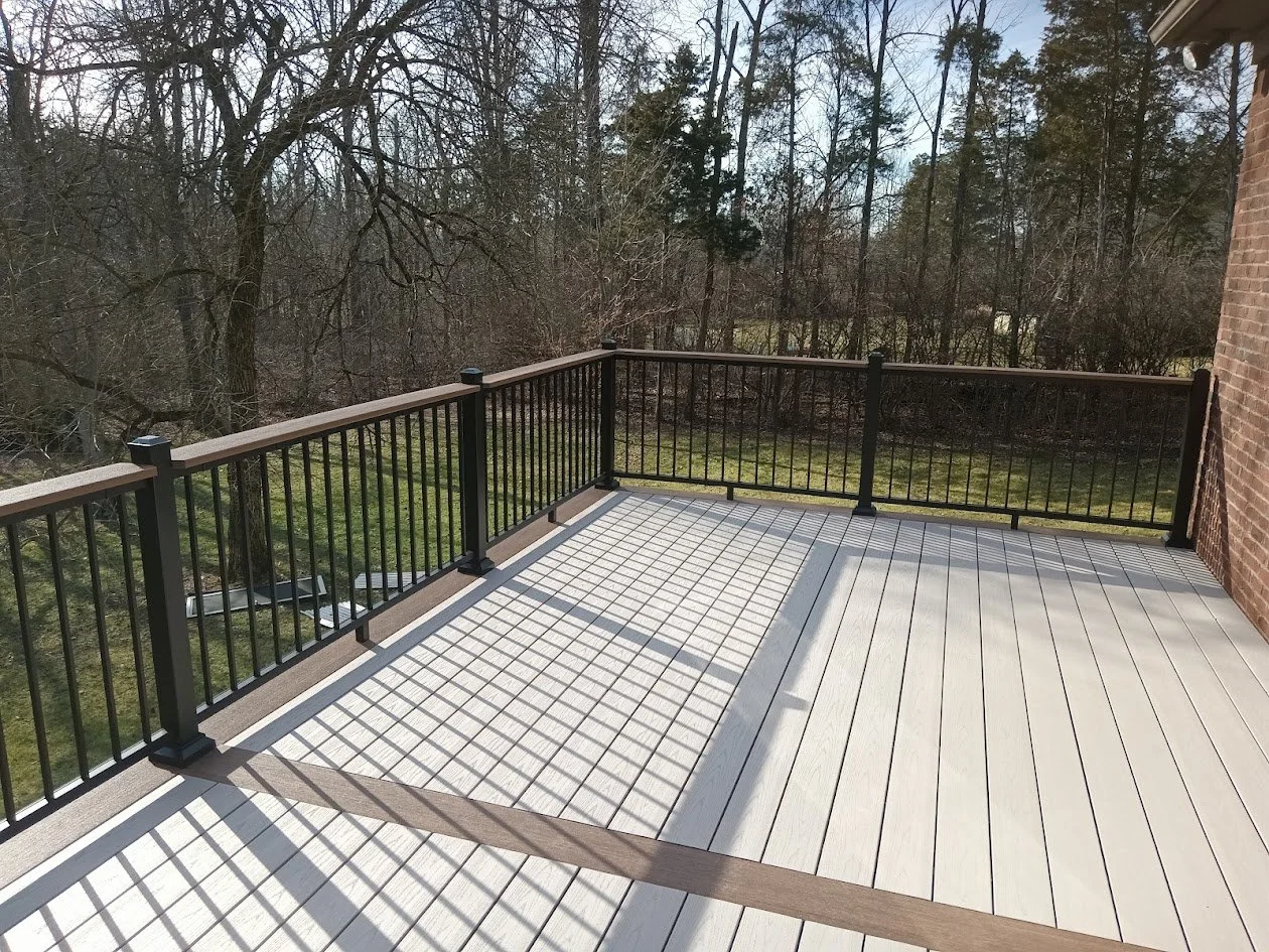 A large outdoor balcony with light-colored wooden flooring, black metal railing, and a view of leafless trees and grass beyond.