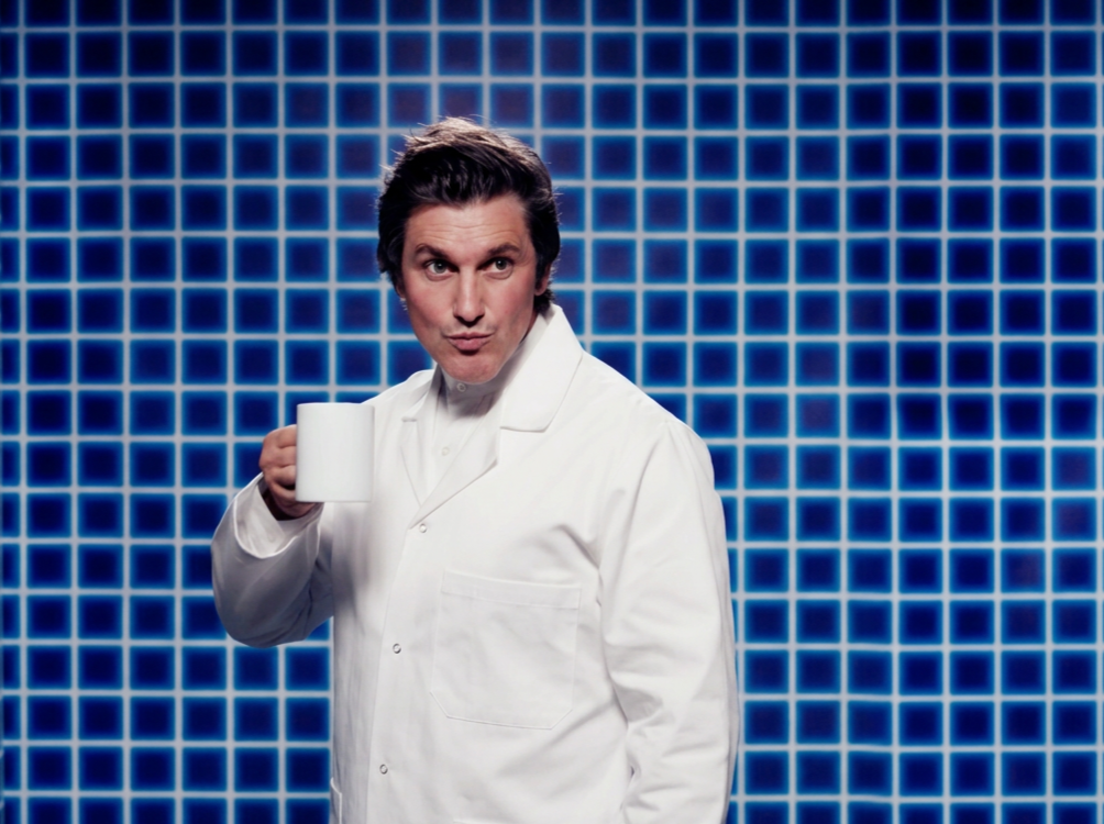 Survivor NZ campaign portrait of the host as a scientist in white lab coat with coffee mug against a blue grid, styled within the campaign’s eccentric pseudo-scientific world.