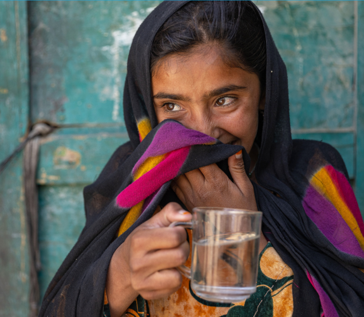 Documentary portrait of a girl holding a glass of clean water, her smile partly covered by fabric, grounding UNICEF’s life-changing work in an individual human reality.
