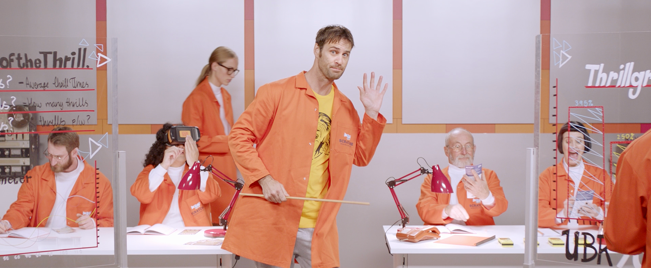 The host moves through a frame of simultaneous foreground and background experiments, using longer staged movement to carry several ideas at once while giving the script’s density room to breathe instead of relying on frenetic cutting.