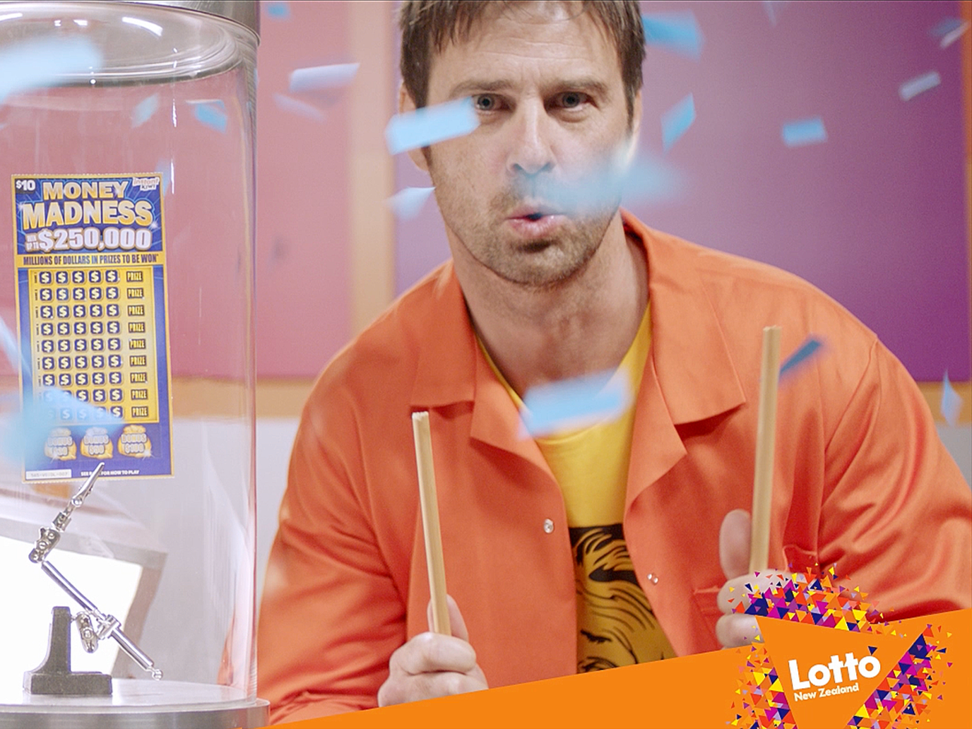 The Instant Kiwi campaign host in research uniform amid falling confetti, combining heightened spectacle with the institute’s straight-faced seriousness so the world remains playful without breaking its internal truth.