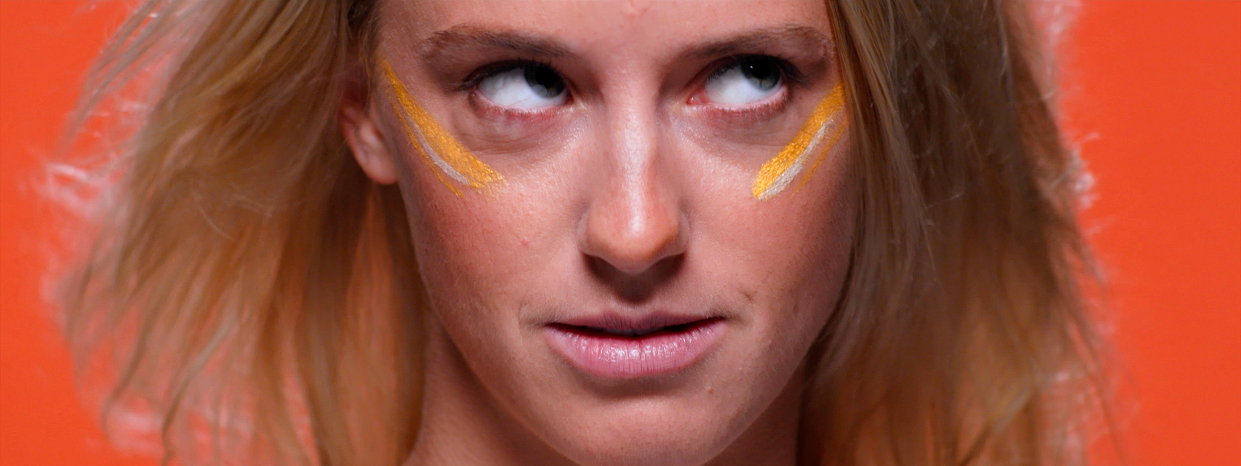 Close-up Survivor NZ campaign portrait of a contestant with orange markings beneath her eyes, continuing the heightened character-study treatment as she rolls her eyes.