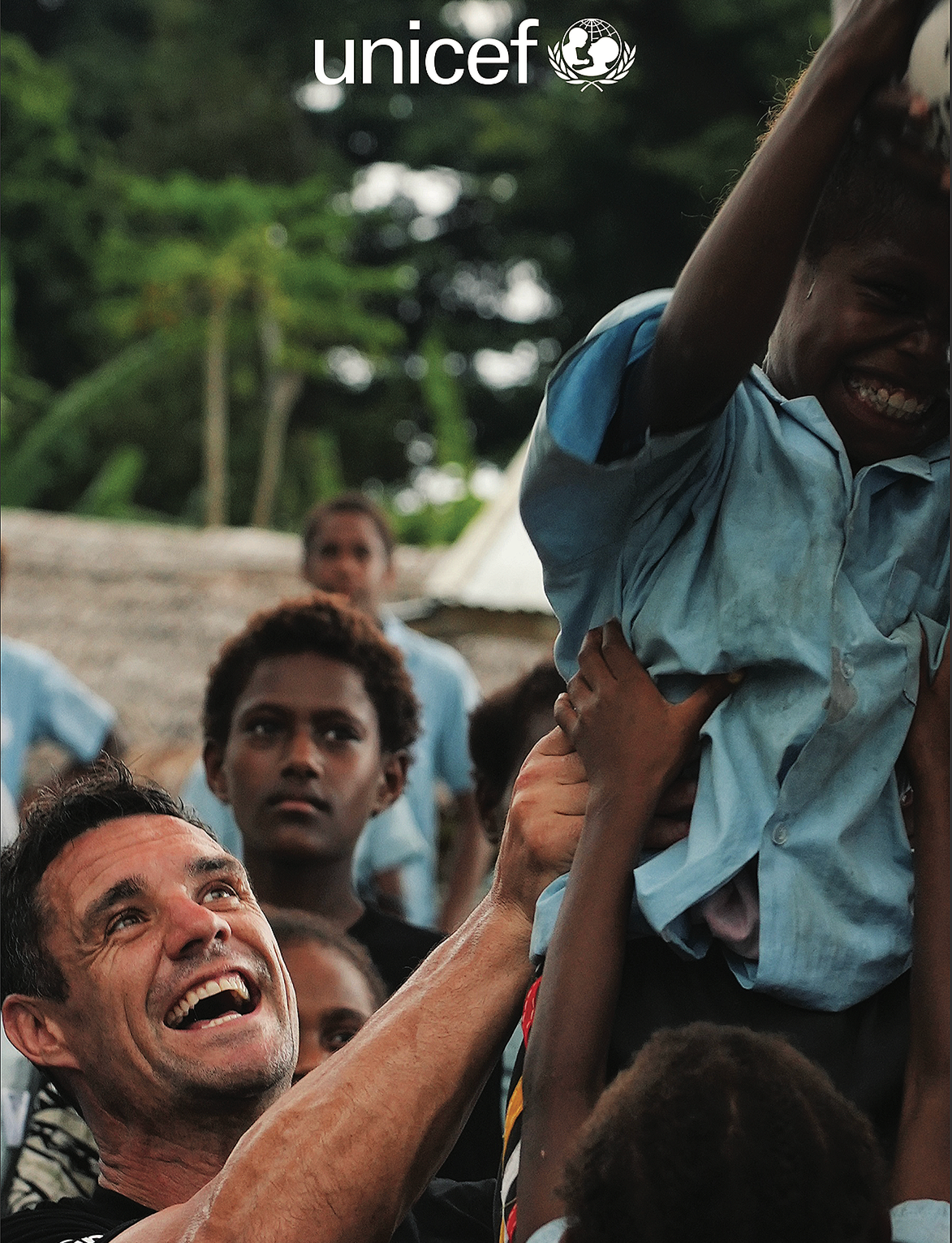 Campaign photograph of Dan Carter lifting a child among a group of children, shifting the visual story from public sporting activity back towards the people and communities the fund exists to support.